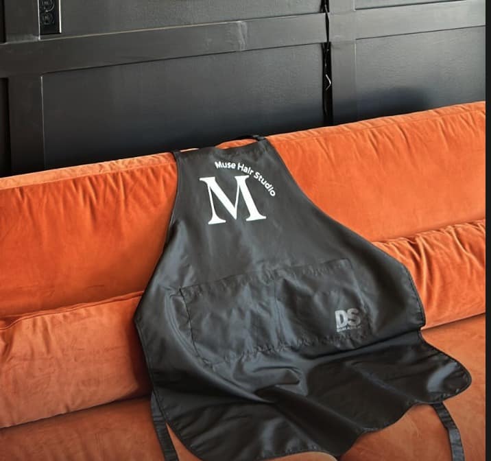 Muse Hair Studio branded apron on velvet couch beneath gold logo signage