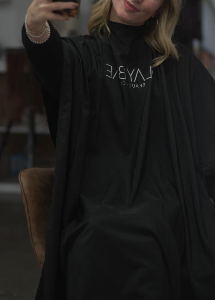 Client in a Slaybae Beauty Co branded salon cape taking a mirror selfie